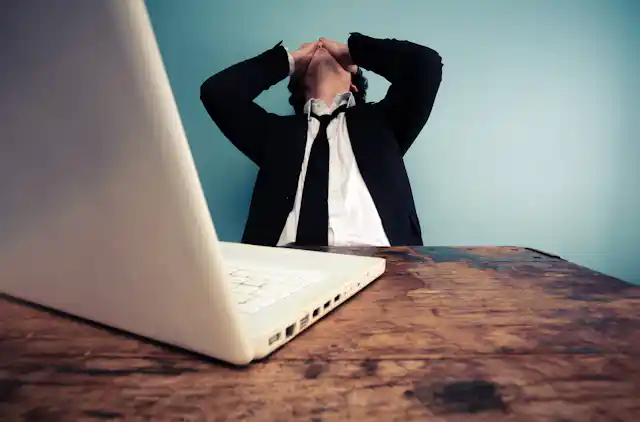 Man in suit, head back, hands covering face, blue background, laptop in front.