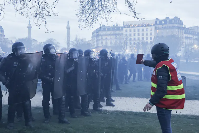 A protestor holding a phone up towards a line of riot police holding shields in a street filled with smoke.