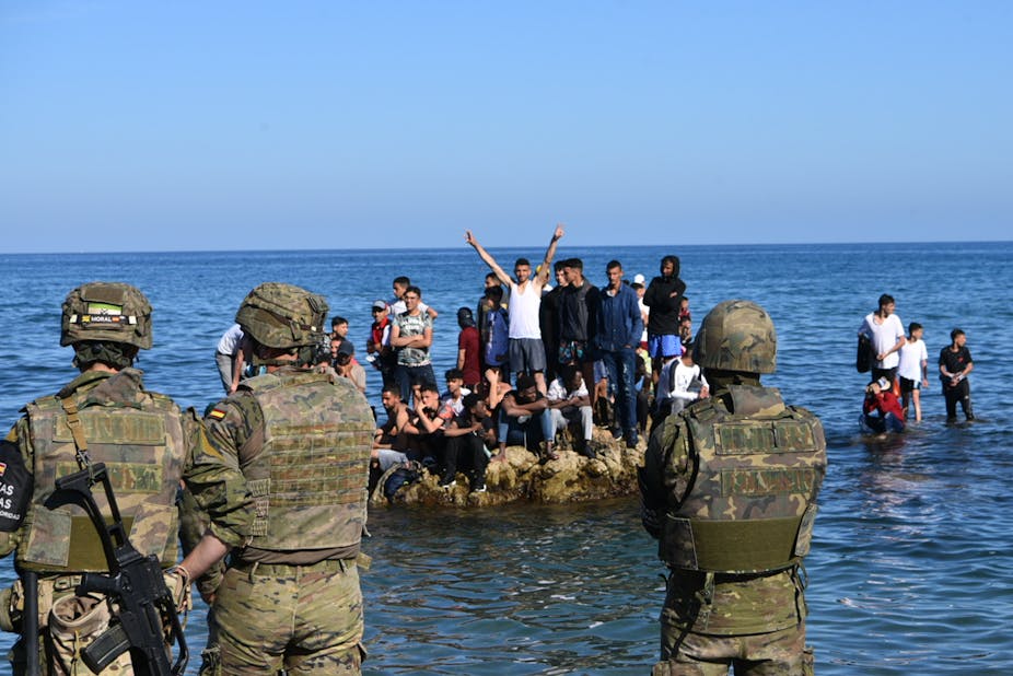 Soldats de dos, faisant face à un groupe de jeunes hommes juchés sur un rocher dans la mer