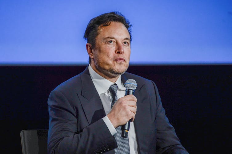 Elon Musk, wearing a suit and tie, holds a microphone and looks up with pursed lips