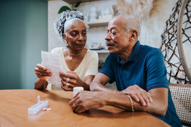 Two people read a package insert together with a weekly pill organizer on the table before them