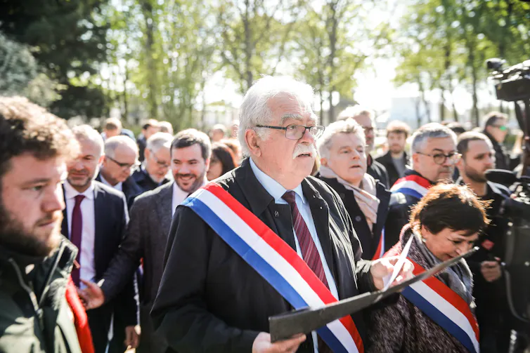 Men and women wearing tricolore sashes walking along a street in a crowd.
