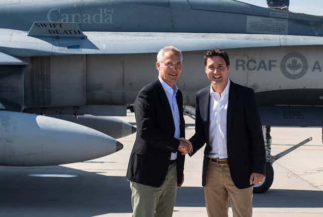 Two men, one with thinning grey hair and glasses, and another with dark hair, smile as they shake hands with a fighter jet on a tarmac behind them.
