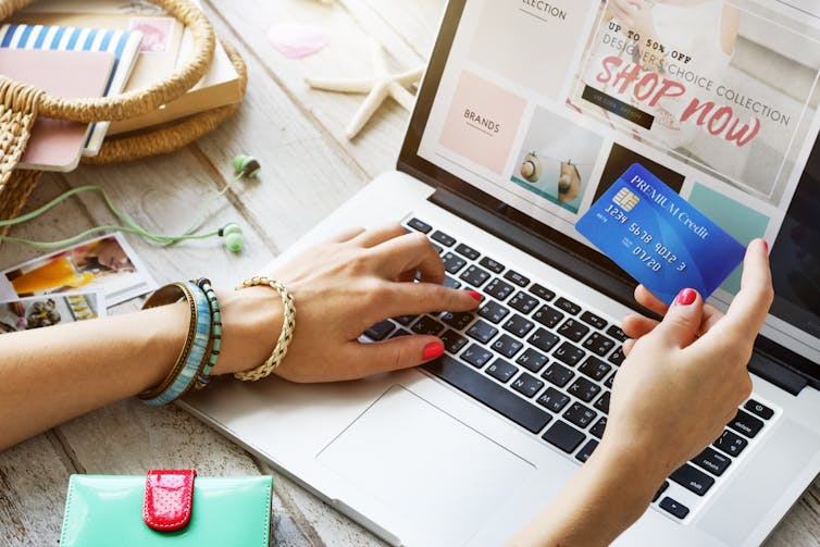 A hand holding a credit card up while another hand navigates an online store on an open laptop.