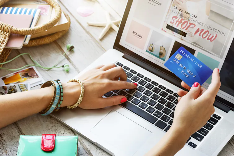 A hand holding a credit card up while another hand navigates an online store on an open laptop.