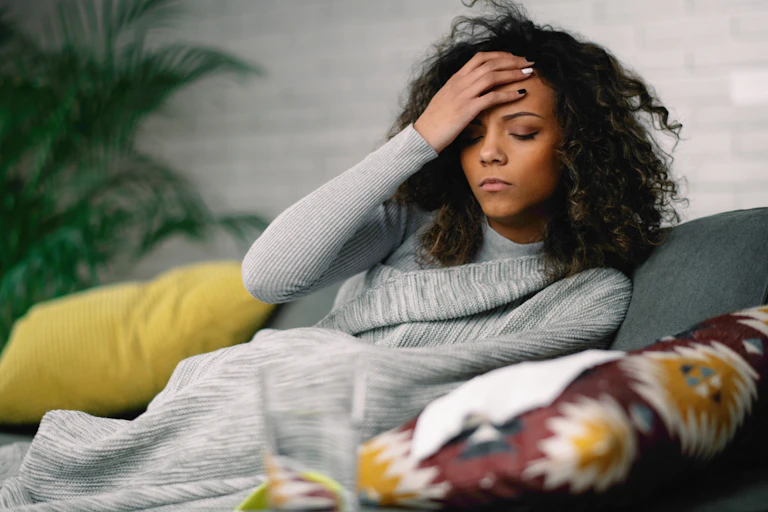 Women on sofa, wrapped in blanket, hand on forehead, eyes shut