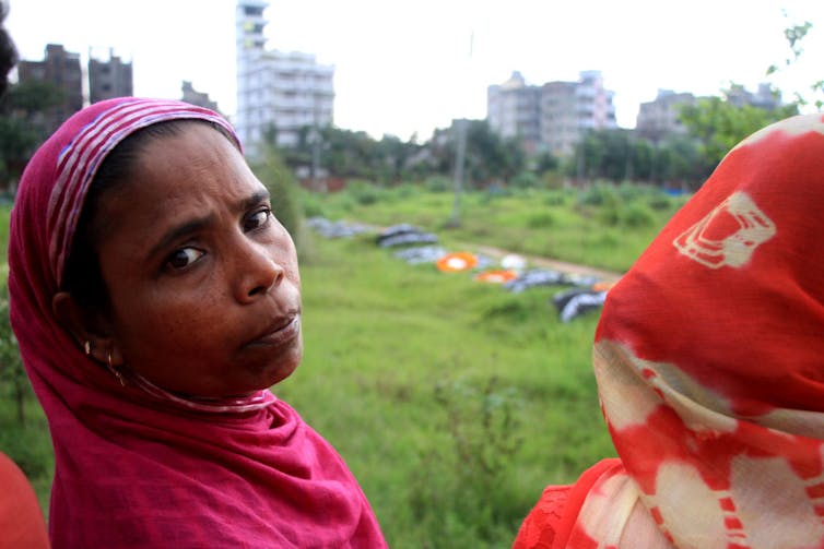 A woman in a pink shawl stares at the camera, with a green field amid tall buildings behind her.