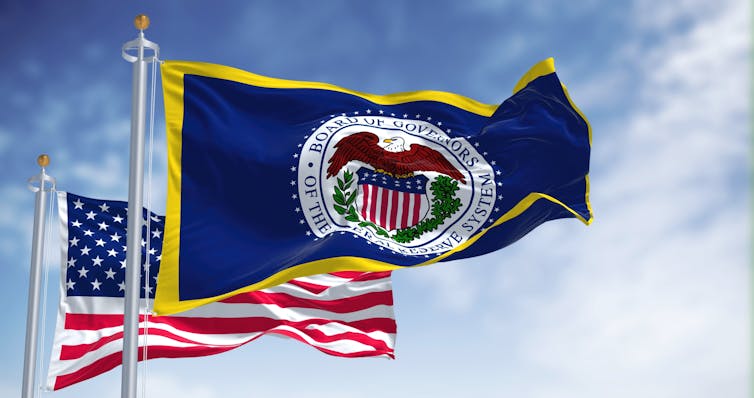 The flag of the US Federal Reserve Board waving in front of the US flag and a blue sky with clouds.