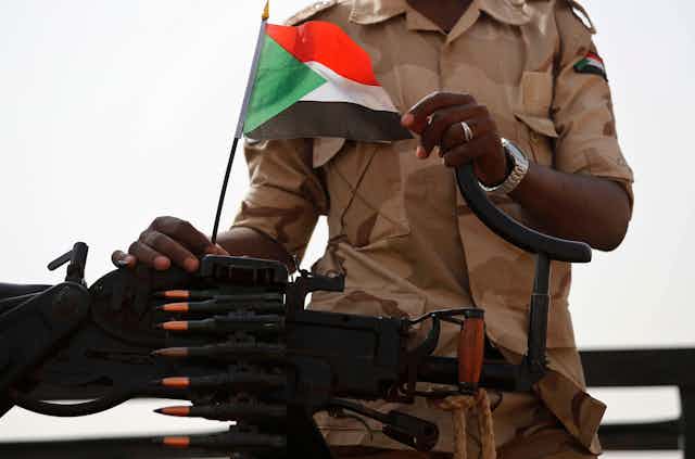 An African soldier from the nexk down holding a machine gun and a Sudan flag.