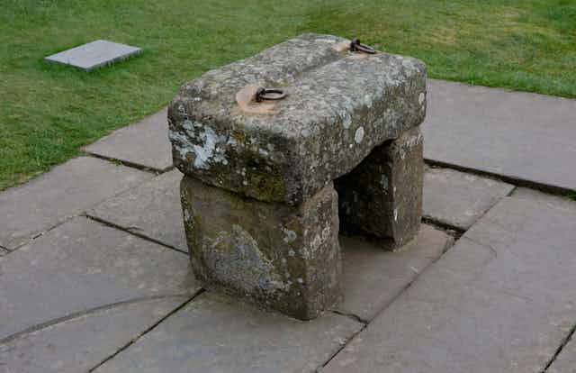 A rectangular slab of stone with two metal rings on top to lift it, rests atop two other slabs of stone to form a bench shape