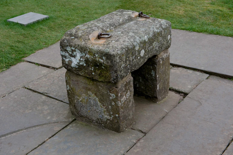 A rectangular slab of stone with two metal rings on top to lift it, rests atop two other slabs of stone to form a bench shape