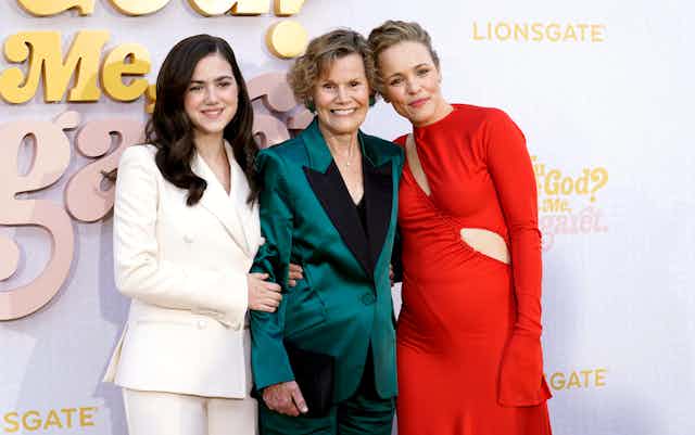 A young woman in a white suit, an older woman in a teal suit and a middle-aged blonde woman in a red dress with a cut-out smile together in front of a sign: Are You There, God? It's me, Margaret