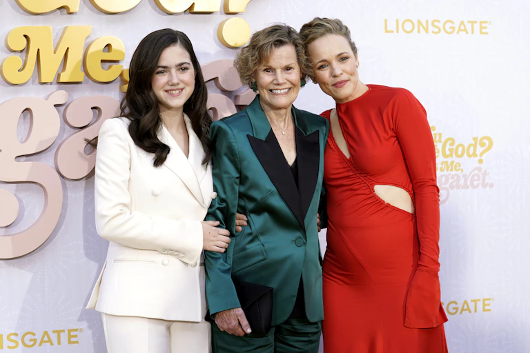 A young woman in a white suit, an older woman in a teal suit and a middle-aged blonde woman in a red dress with a cut-out smile together in front of a sign: Are You There, God? It's me, Margaret