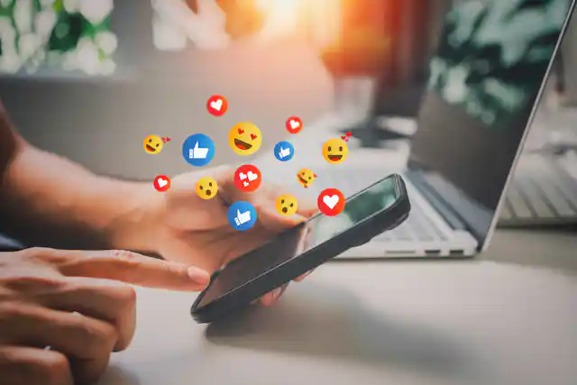 Person scrolls on phone while heart, thumbs up and other emojis float above screen