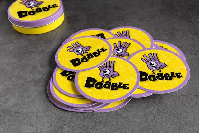 A pile of yellow and purple discs piled up on a grey background. The word 'Dobble' is written on each disc alongside a cartoon character.