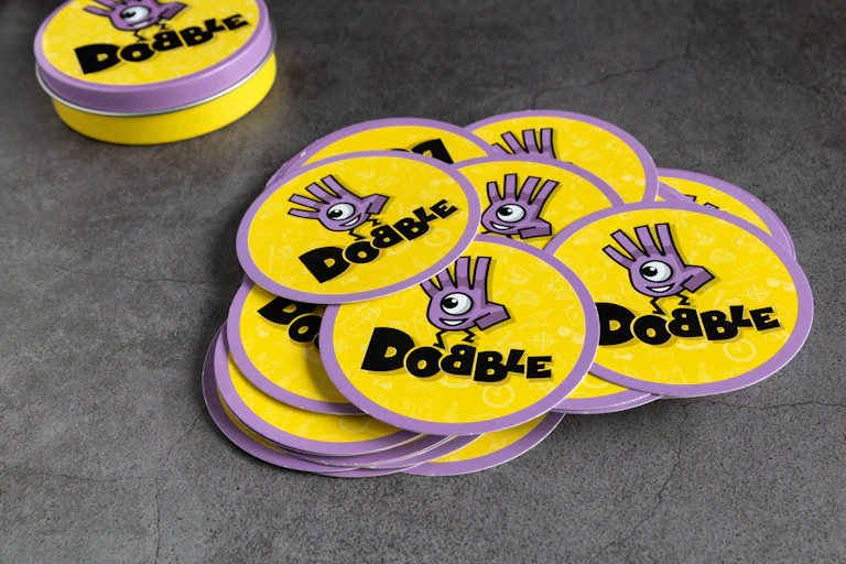 A pile of yellow and purple discs piled up on a grey background. The word 'Dobble' is written on each disc alongside a cartoon character.