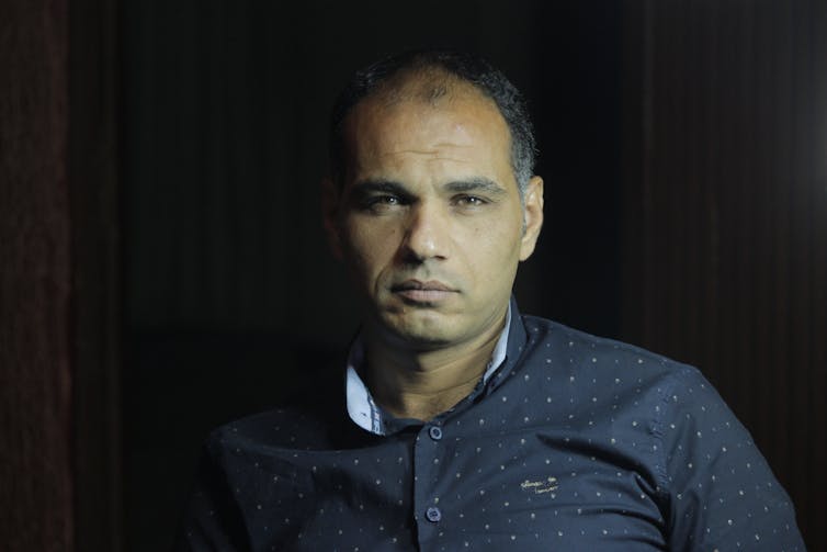 A brown-skinned middle-aged man looks directly at the camera. He wears a dark blue shirt and sits in front of a dark background.