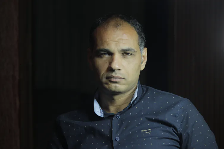 A brown-skinned middle-aged man looks directly at the camera. He wears a dark blue shirt and sits in front of a dark background.