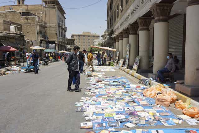 A bunch of books are laid out on a street. People stand over the books and look at them. There are grey buildings around the open book market. 