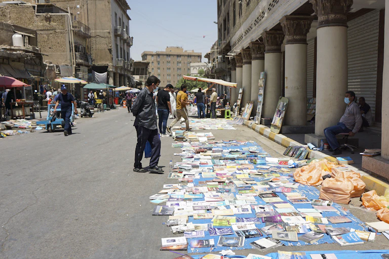 A bunch of books are laid out on a street. People stand over the books and look at them. There are grey buildings around the open book market.