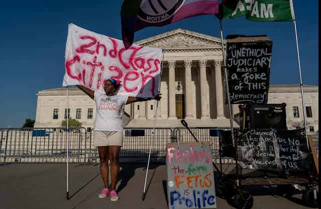 An activist displaying multiple protest posters stands in front of the Supreme Court building.