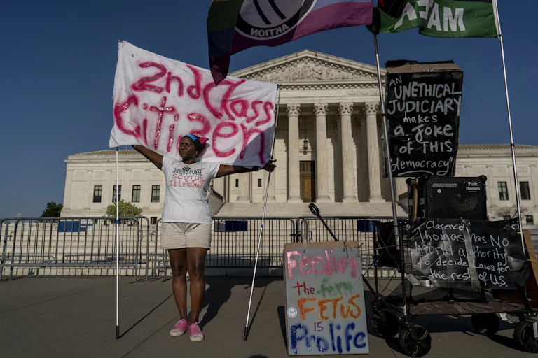 An activist displaying multiple protest posters stands in front of the Supreme Court building.