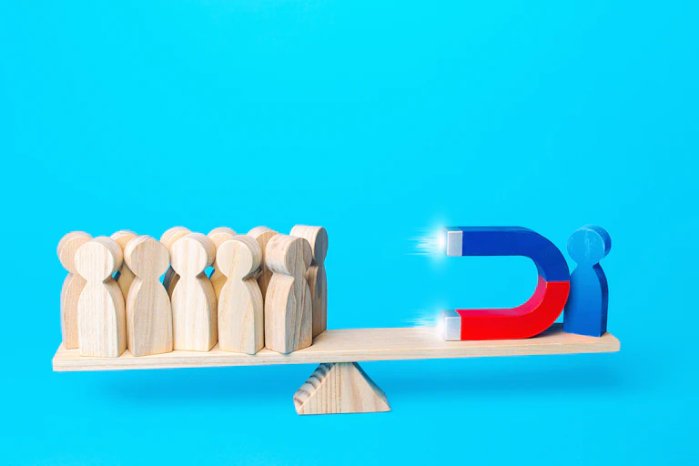 A blue wooden figure with a huge magnet, balanced on one side of a balance beam, tries to attract other wooden figures from the other end of the beam.
