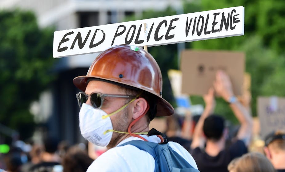A man wearing a brown construction hat, sunglasses and a mask over his nose and mouth holds a sign that reads "End Police Violence" during a protest.