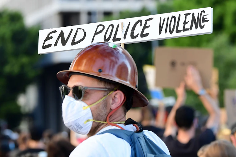 A man wearing a brown construction hat, sunglasses and a mask over his nose and mouth holds a sign that reads "End Police Violence" during a protest.