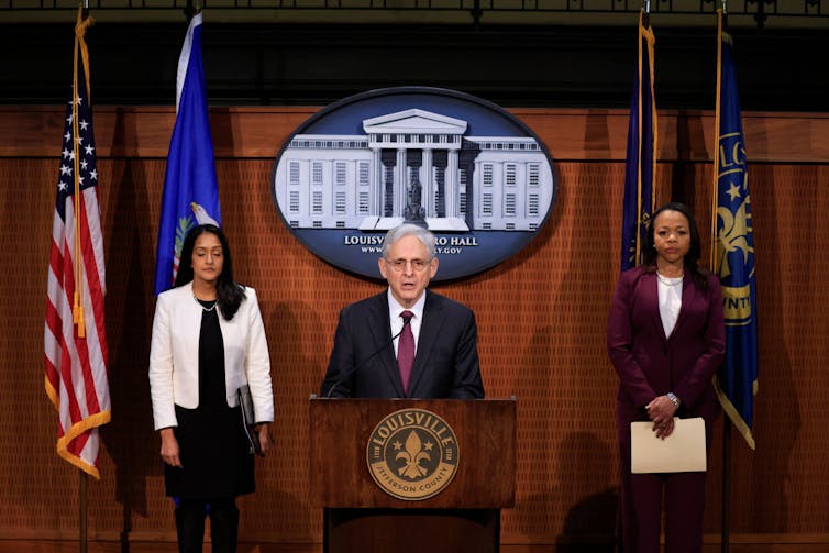 Flanked by two suited women, a gray-haired man in a suit and wearing glasses speaks behind a lectern with the emblem 