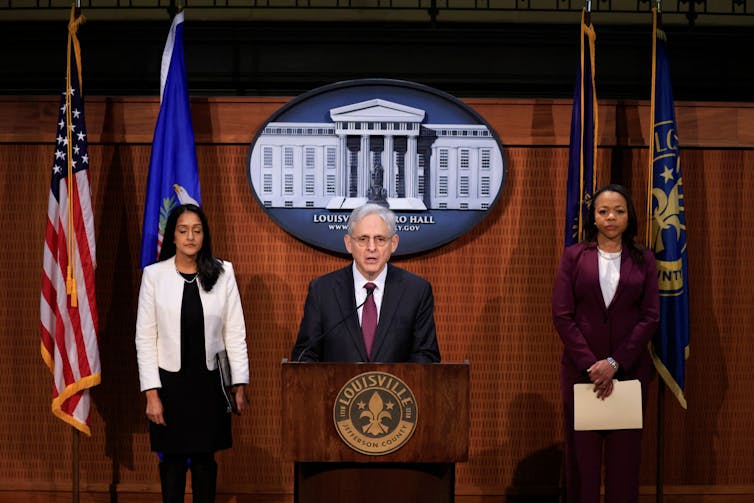 Flanked by two suited women, a gray-haired man in a suit and wearing glasses speaks behind a lectern with the emblem
