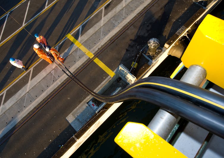 2 long, thick electric cables are lowered from a ship to workers on the dock below.