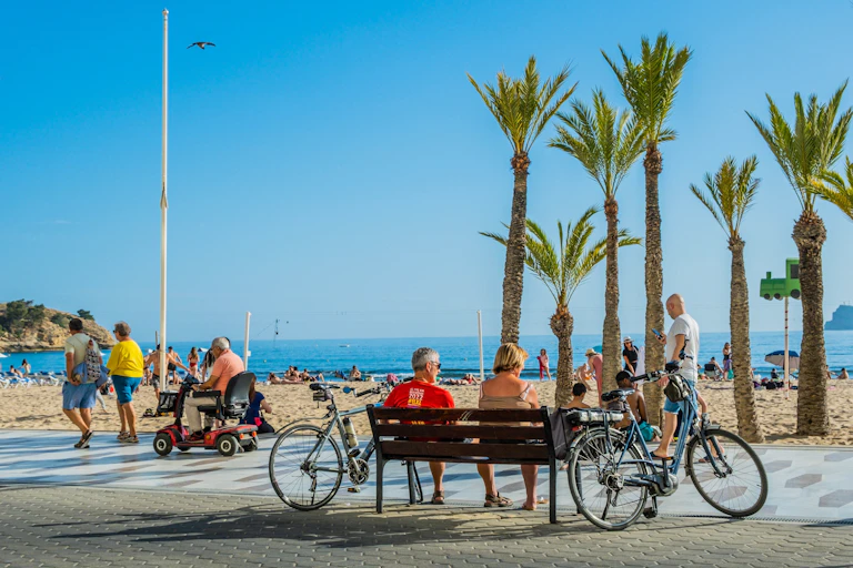 Personas paseando por el paseo marítima frente a una playa con gente.
