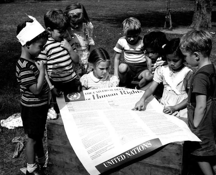 A black and white photo shows a handful of children studying a large poster with small text printed on it.