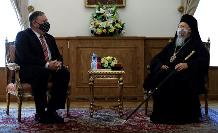 Two men in face masks – one in a suit, another in black clerical garb – sit formally during a meeting.