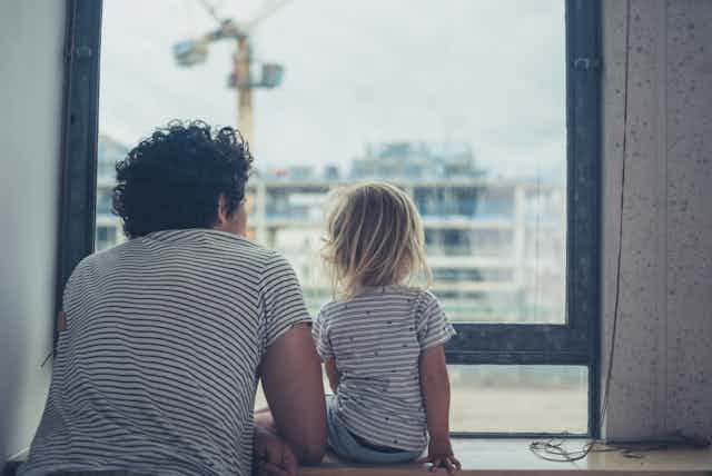 Father and small daughter look out of apartment window at another apartment under construction
