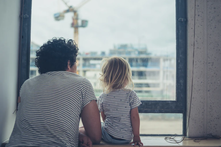 Father and small daughter look out of apartment window at another apartment under construction