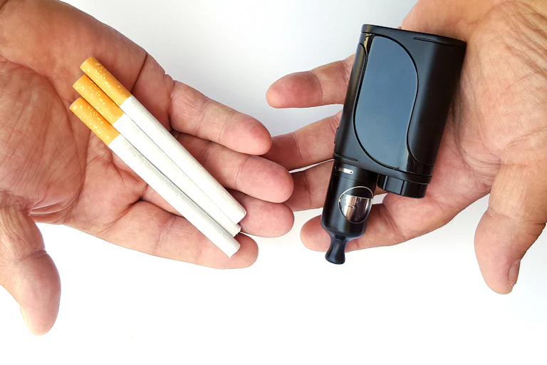 A person holds three cigarettes in one hand and a disposable vape in the other hand.