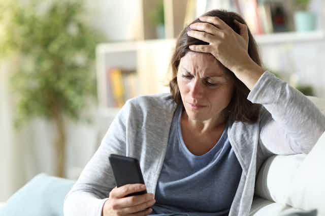 A middle aged woman sits on a couch looking at her mobile phone with her hand on her forehead and a confused expression on her face
