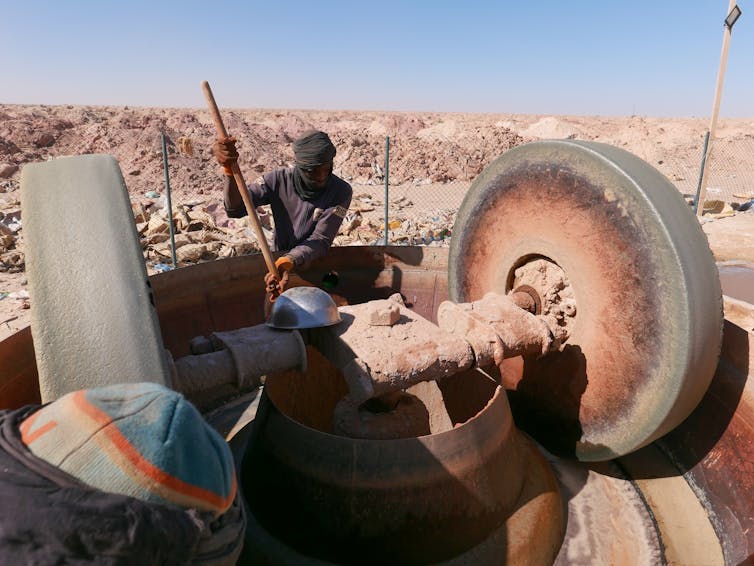 A person wearing a head scarf and holding a shovel operates a machine crushing ore.