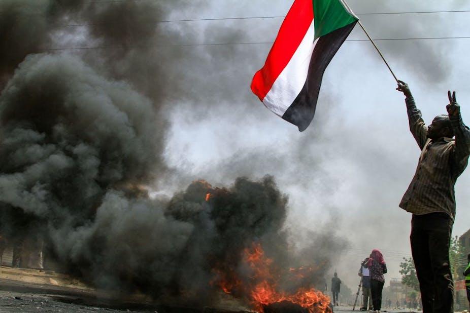 A man holding up a flag in one hand while the other hand shows the two-finger peace sign stands near heavy smoke created by a burning tyre.