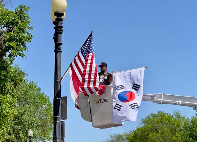 A man in a baseball cap in a crane fixes an American flag and a South Korean flag to a lamp post.