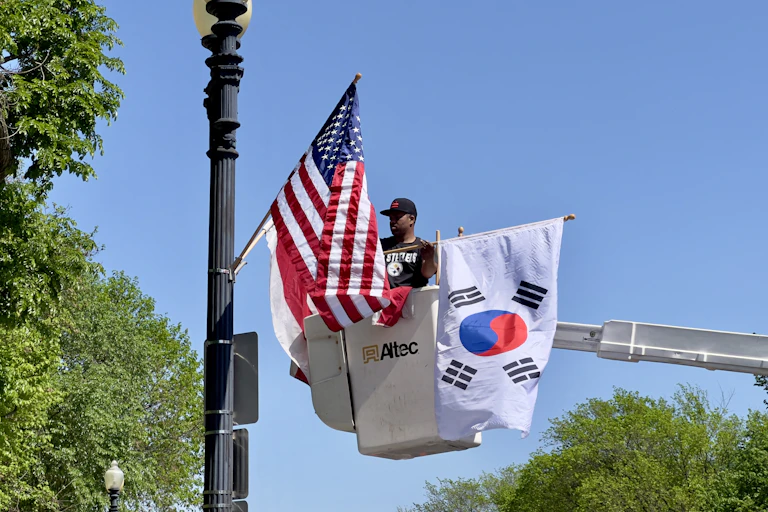 A man in a baseball cap in a crane fixes an American flag and a South Korean flag to a lamp post.