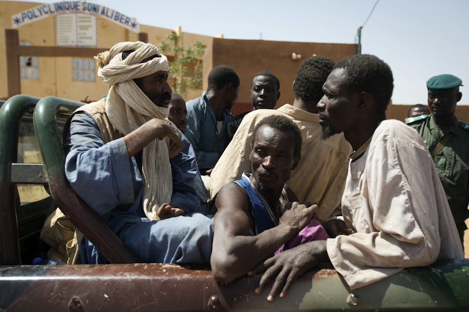 Six hommes assis dans l'arrière d'un véhicule sous le regard d'un individu en uniforme