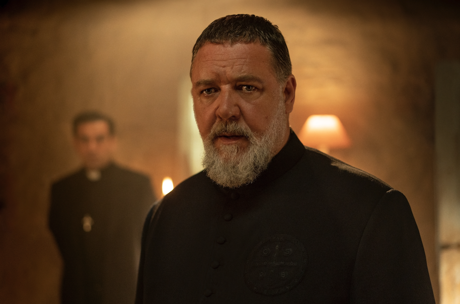 Russell Crowe in all black, with a priest stood behind him. 