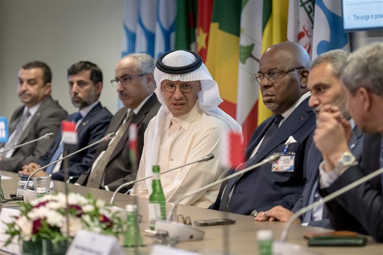 Panel of OPEC ministers in front of a row of flags, desks & microphones in front.