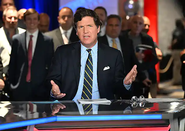 A man in a blazer, blue checked shirt and rep tie, sitting at a table and gesturing with his hands.