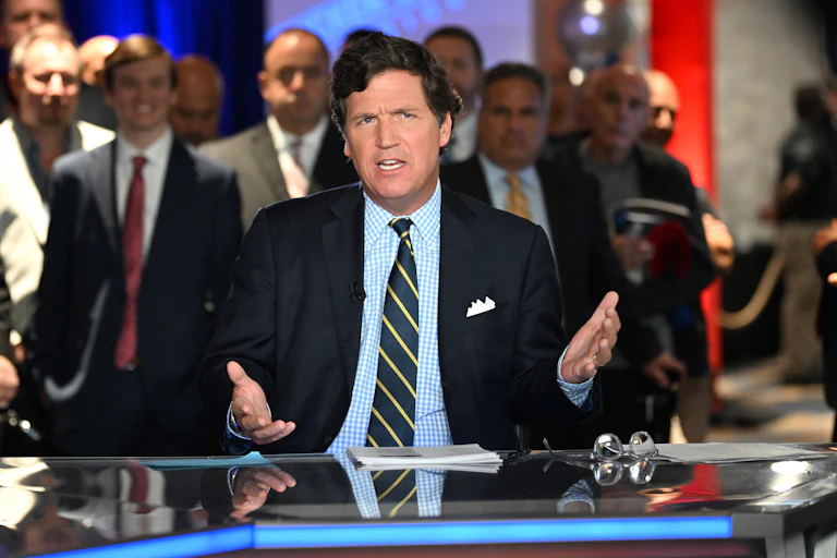A man in a blazer, blue checked shirt and rep tie, sitting at a table and gesturing with his hands.