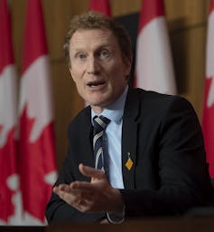 A man in a suit sitting in front of a Canadian flag.