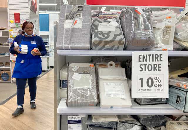 a black woman wearing a blue bed bath and beyond apron holds some products as she walks through aisles at a store, where a sign near bedding materials reads entire store 10% off